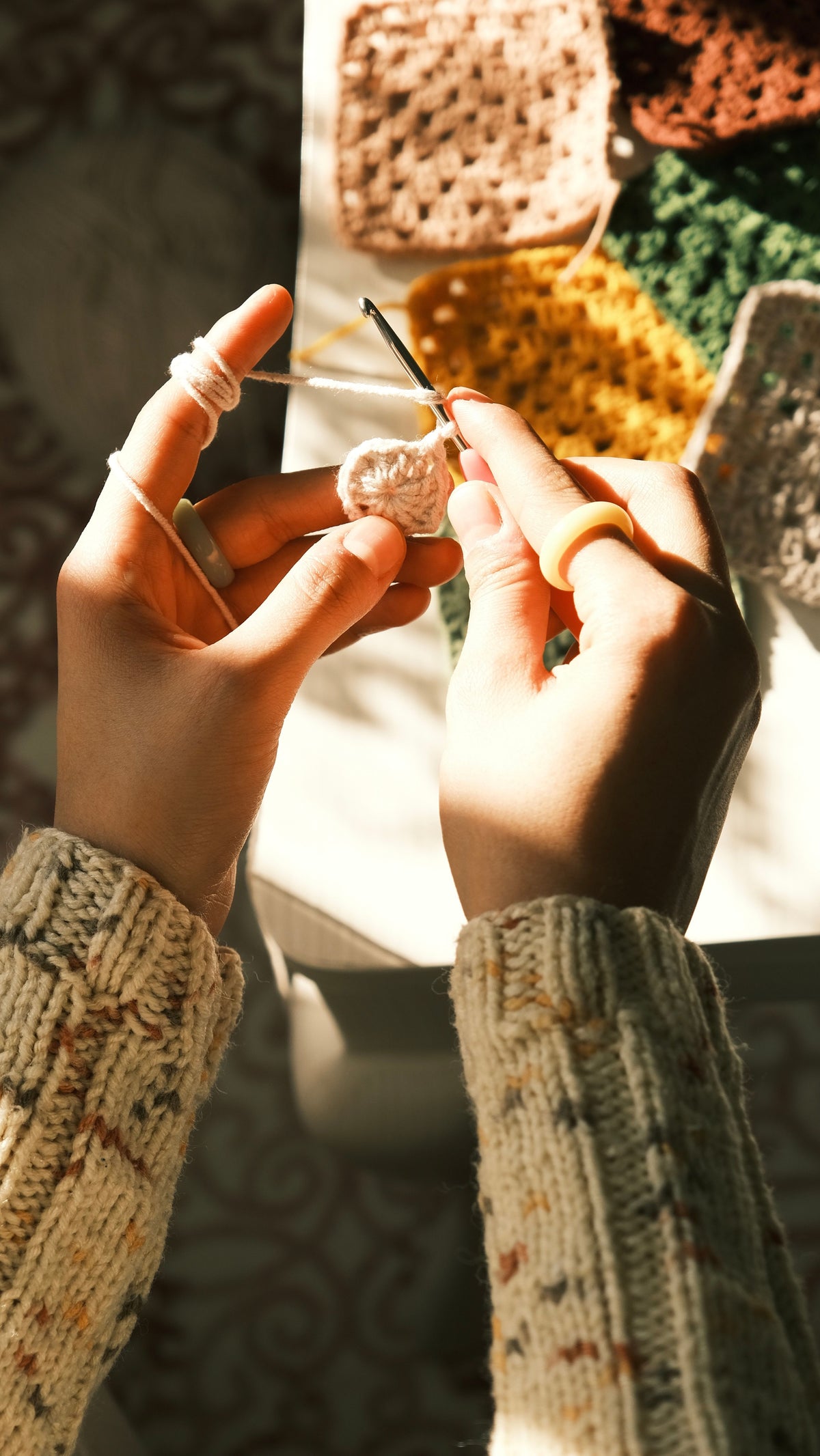 Hands crocheting a granny square in warm sunlight, with colorful crochet squares in the background